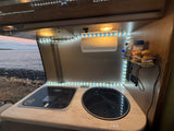 An RV rental kitchen with a stovetop, sink, and view of the ocean, illuminated by LED lights.