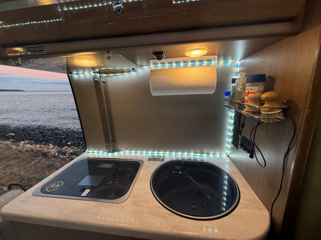 An RV rental kitchen with a stovetop, sink, and view of the ocean, illuminated by LED lights.