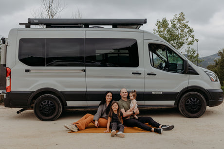 Smiling family of four on a blanket by a white camper van, ideal for an RV rental outdoor adventure.