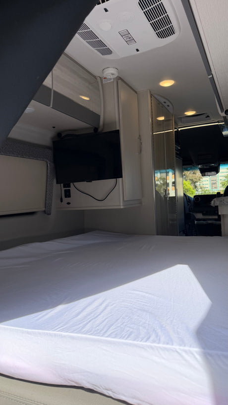 Relax in this rv rental! Modern camper van interior with a bed, mounted TV, and overhead AC unit. Sunlit sleeping area.