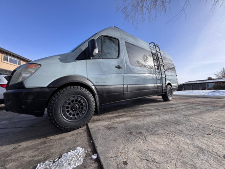 Light grey activan camper van with off-road tires & ladder, parked on a sunny driveway. Perfect rv rental adventure awaits!