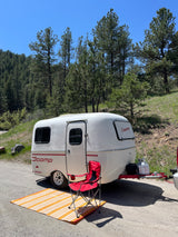 Scamp travel trailer for your next RV rental adventure. Parked in a mountain forest setting with a camping chair and mat.
