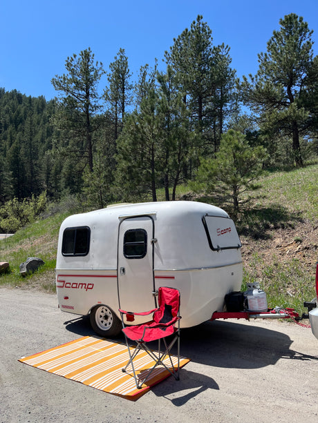 Scamp travel trailer for your next RV rental adventure. Parked in a mountain forest setting with a camping chair and mat.