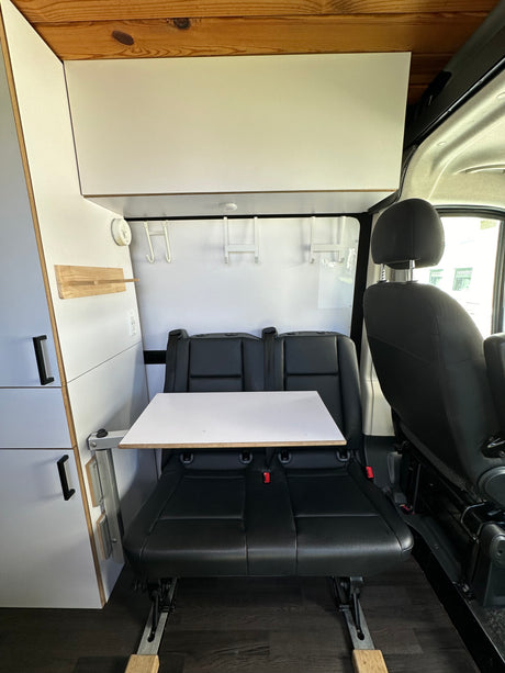 Modern RV rental interior with dual black leather seats, a flexible white table, and integrated white storage cabinets.