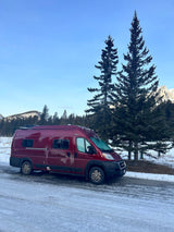 Maroon rv rental camper van on snowy road. Tall evergreens & snowy mountains under blue sky for winter adventure.