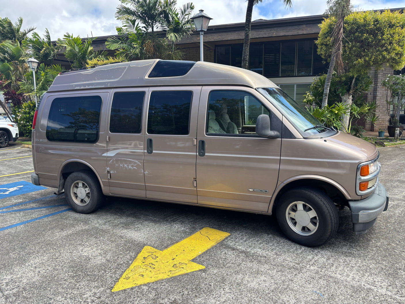 Tan Chevrolet conversion van, used for rv rental, parked in a lot with palm trees. Features a raised roof & paint wear.