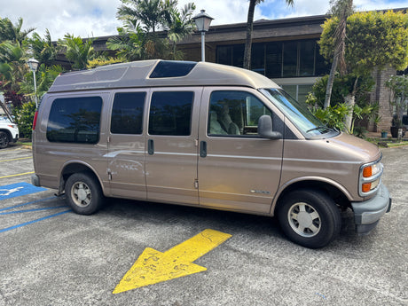 Tan Chevrolet conversion van, used for rv rental, parked in a lot with palm trees. Features a raised roof & paint wear.