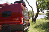 Rear view of a red Solis RV rental camper van with Coleman Mach Q AC, parked lakeside in a grassy, wooded area.