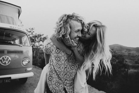 A smiling couple shares a joyful piggyback, with a vintage VW camper in the background, perfect for an RV rental adventure.