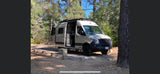 Experience the outdoors in this beige Mercedes Sprinter **rv rental** parked at a wooded campsite with its doors open.