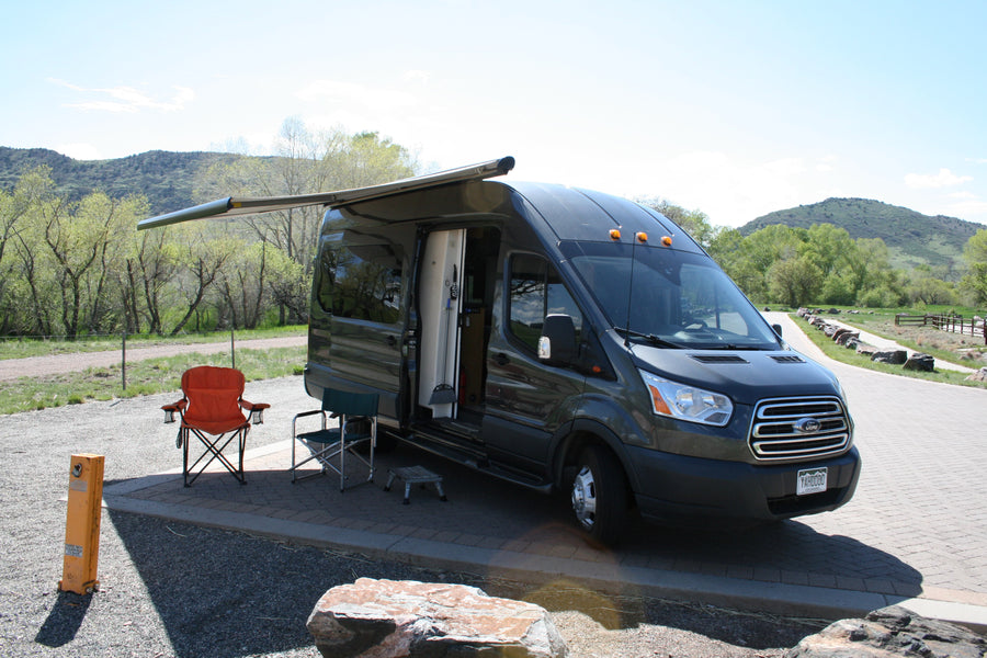 Gray Ford Transit RV rental camper van with extended awning & chairs at a scenic mountain campsite, ready for outdoor travel.