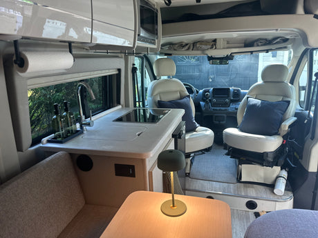 Clean RV rental interior: kitchen with sink & cooktop, dining table with lamp, and front cabin with swivel seats.