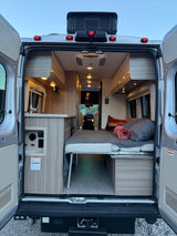 Interior of an RV rental van showing the bed, kitchen, and open rear doors. Coleman Mach AC unit is visible.