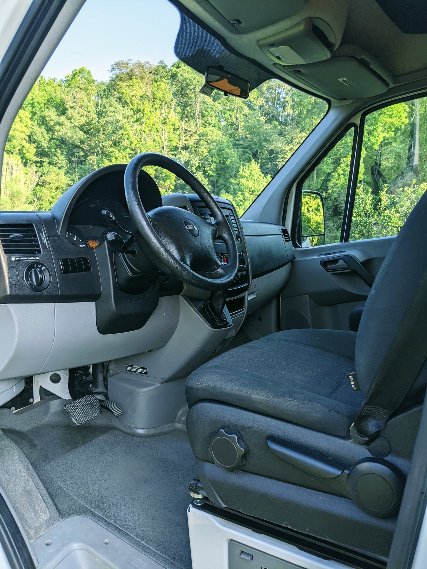 RV rental van interior: Driver's seat, Freightliner dashboard, and a clear view of green trees through the windshield.