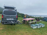 An RV rental camper van parked at an oceanfront campsite. A person sits at a picnic table; bikes are nearby.