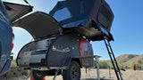 This overland rv rental features a black teardrop trailer with an open galley kitchen, rooftop tent, and ladder deployed at a desert campsite.