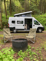 White Winnebago Solis camper van RV rental parked in a lush forest campsite with chairs and fire pit.