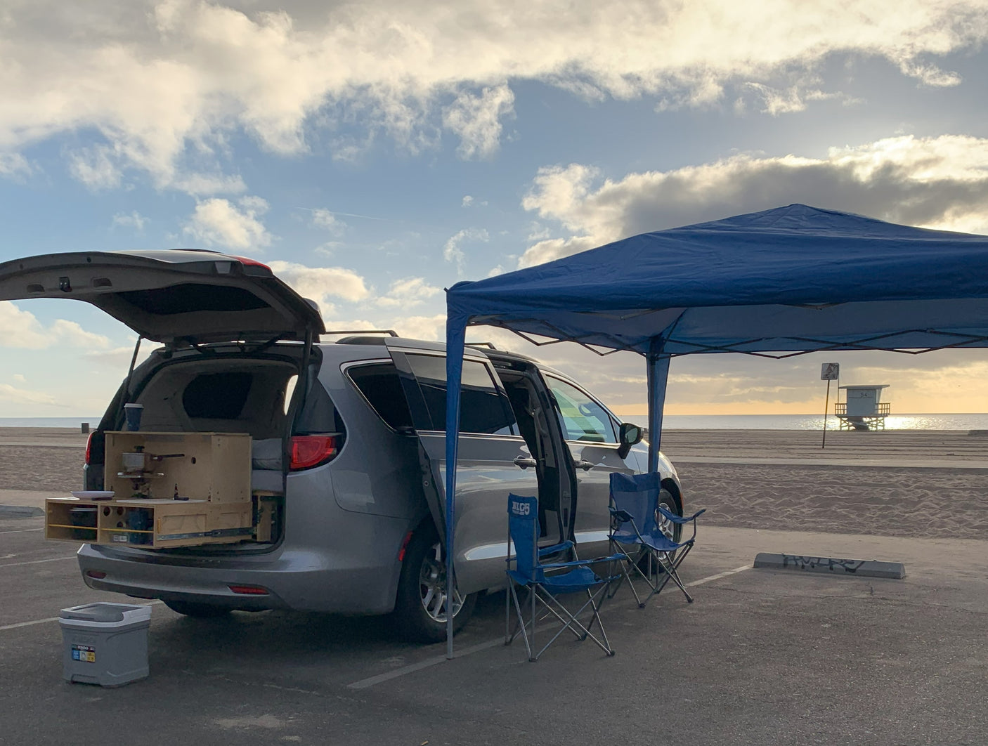 This minivan RV rental is equipped for beach outings: custom kitchen, blue canopy, chairs. Ocean views, lifeguard tower in the distance.