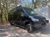 Black Mercedes-Benz Sprinter van RV rental, equipped with an awning & off-road tires, parked amidst autumn trees.