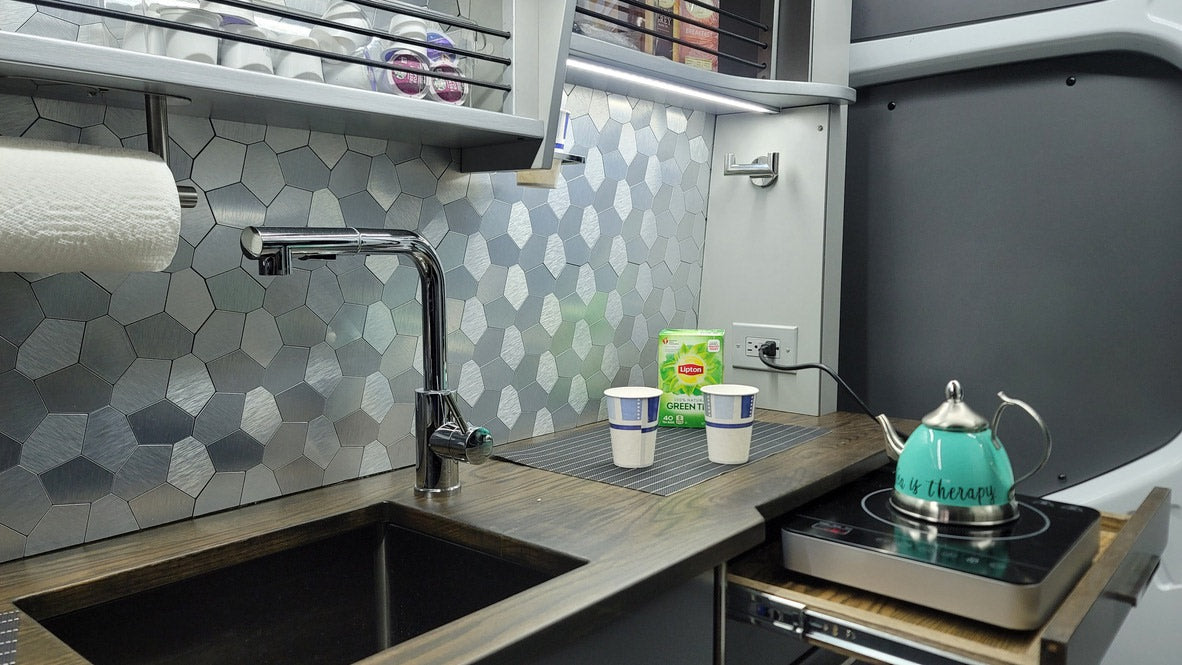 Compact RV rental kitchen features a sleek sink, metallic backsplash, and a teal teapot on an induction cooktop.