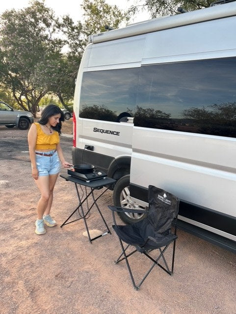 Woman cooking beside her silver Sequence RV rental. She uses a portable stove next to a folding chair for an outdoor meal.