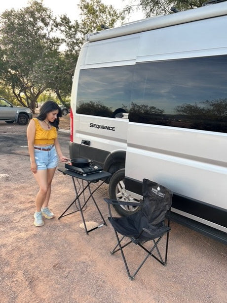 Woman cooking beside her silver Sequence RV rental. She uses a portable stove next to a folding chair for an outdoor meal.