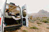 This RV rental offers a cozy escape: a woman reads in her camper van, parked amidst a vast desert and mountains.