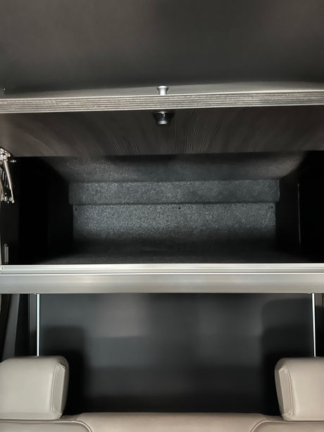 Open overhead storage in an RV rental, revealing a dark, lined interior above light-colored passenger seats.