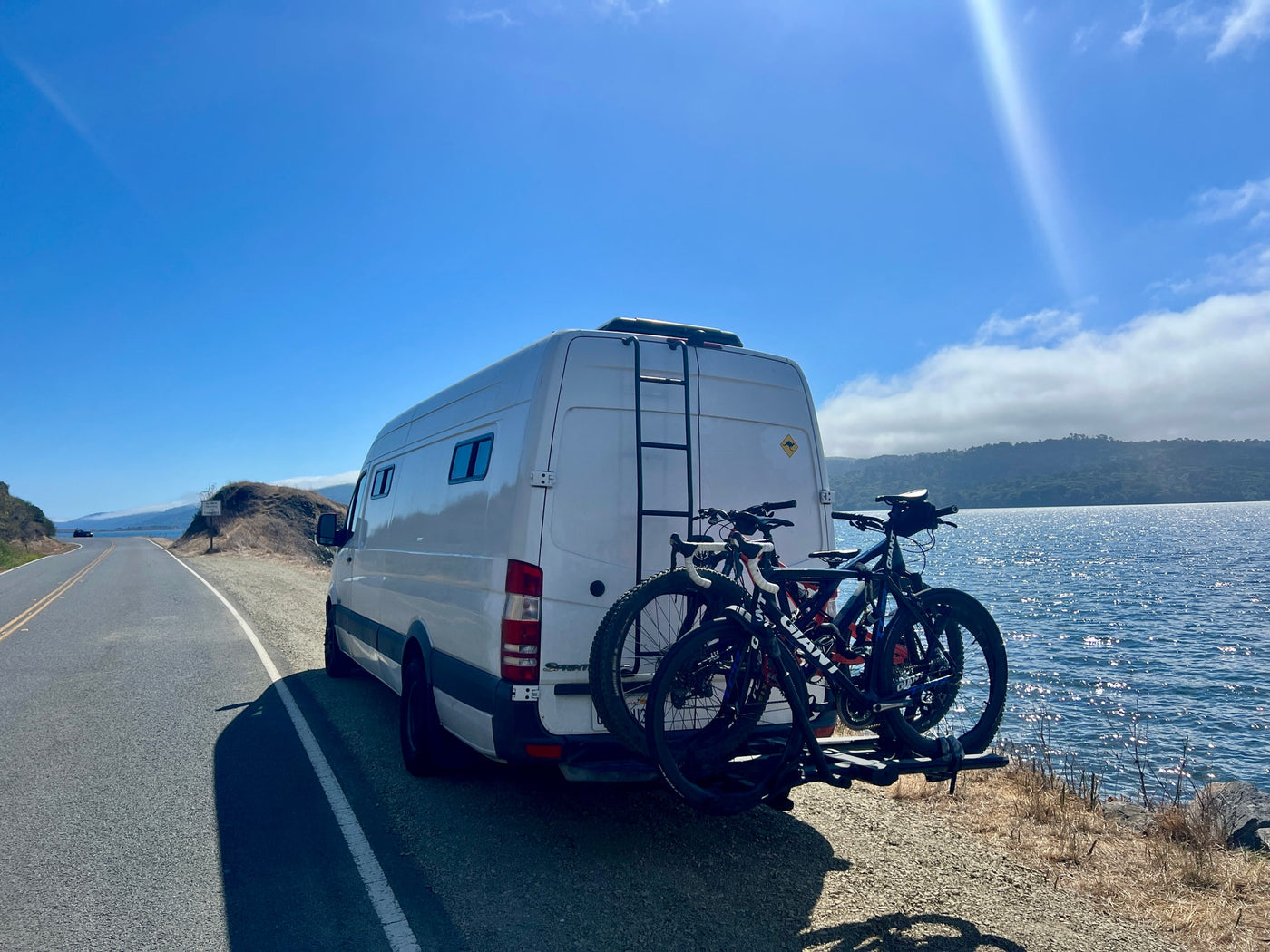 A white Sprinter van, an RV rental, with bikes on the back, parked on a road next to sparkling water on a sunny day. — photo 10
