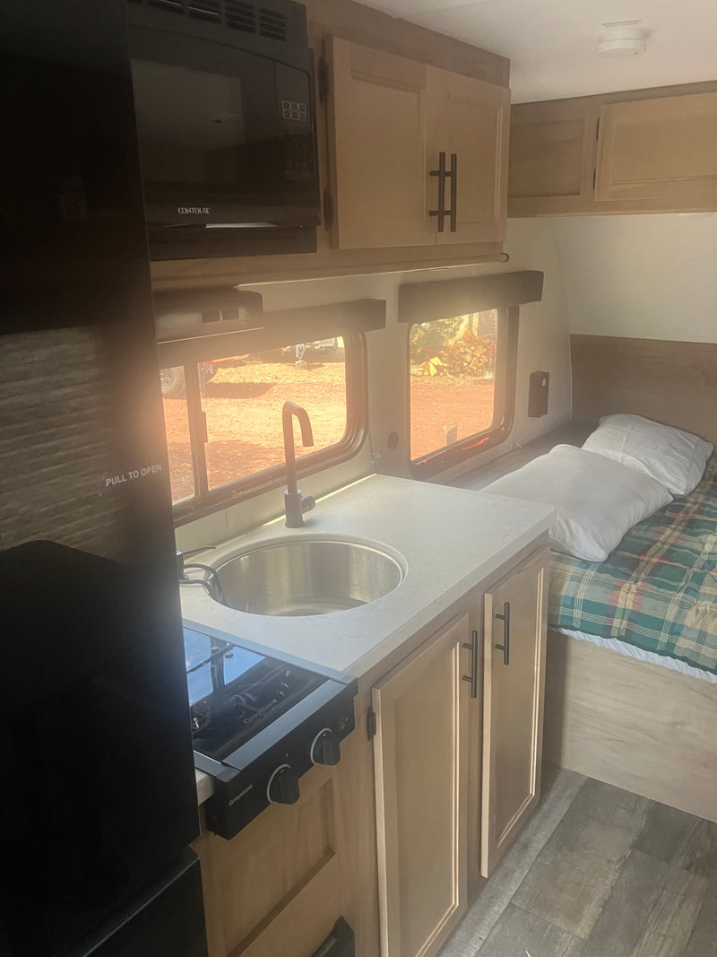 RV rental interior: kitchen with sink, stove, microwave; bed with plaid blanket. Sunny outdoor view through windows. — photo 7