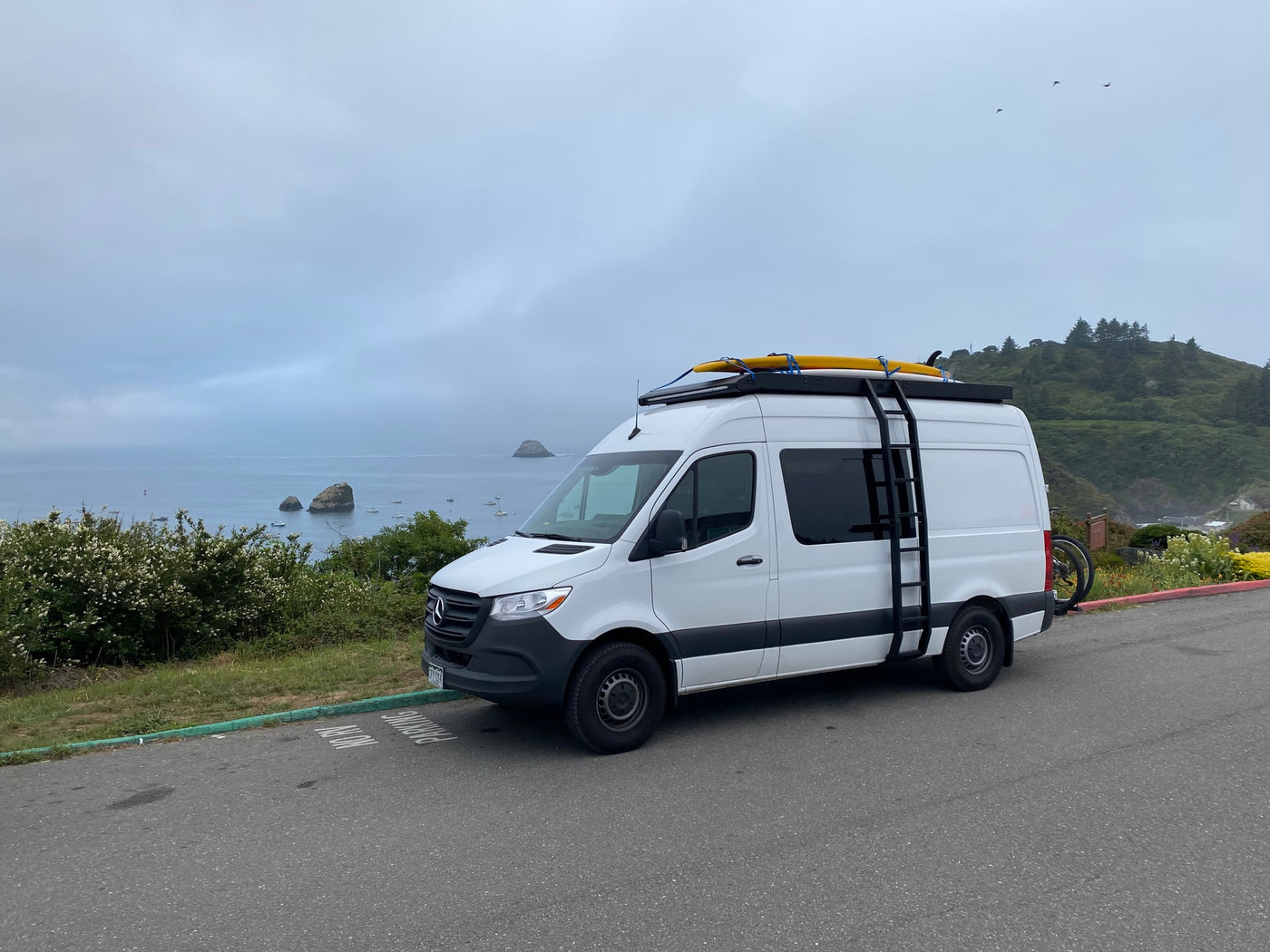 White RV rental van with surfboards & bike overlooking the ocean, rocky islets, and boats on a misty day. — photo 3