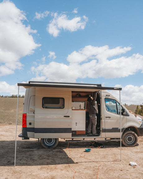 An rv rental camper van parked outdoors with its awning extended. A person is inside the open door, a dog rests on the ground.