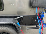 RV rental: Outdoor shower system with blue/red hoses, coiled hose, storage box, and tire on a grey/black trailer.