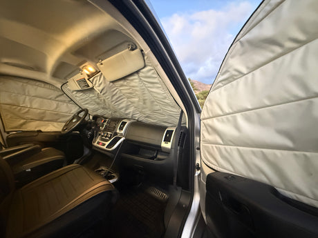 Interior of an RV rental with light-colored quilted window covers, dashboard, and open door showing a mountain view.