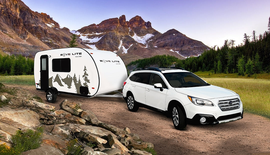 White Subaru Outback tows a Rove Lite teardrop RV rental on a dirt road in mountains & forest, ready for adventure.