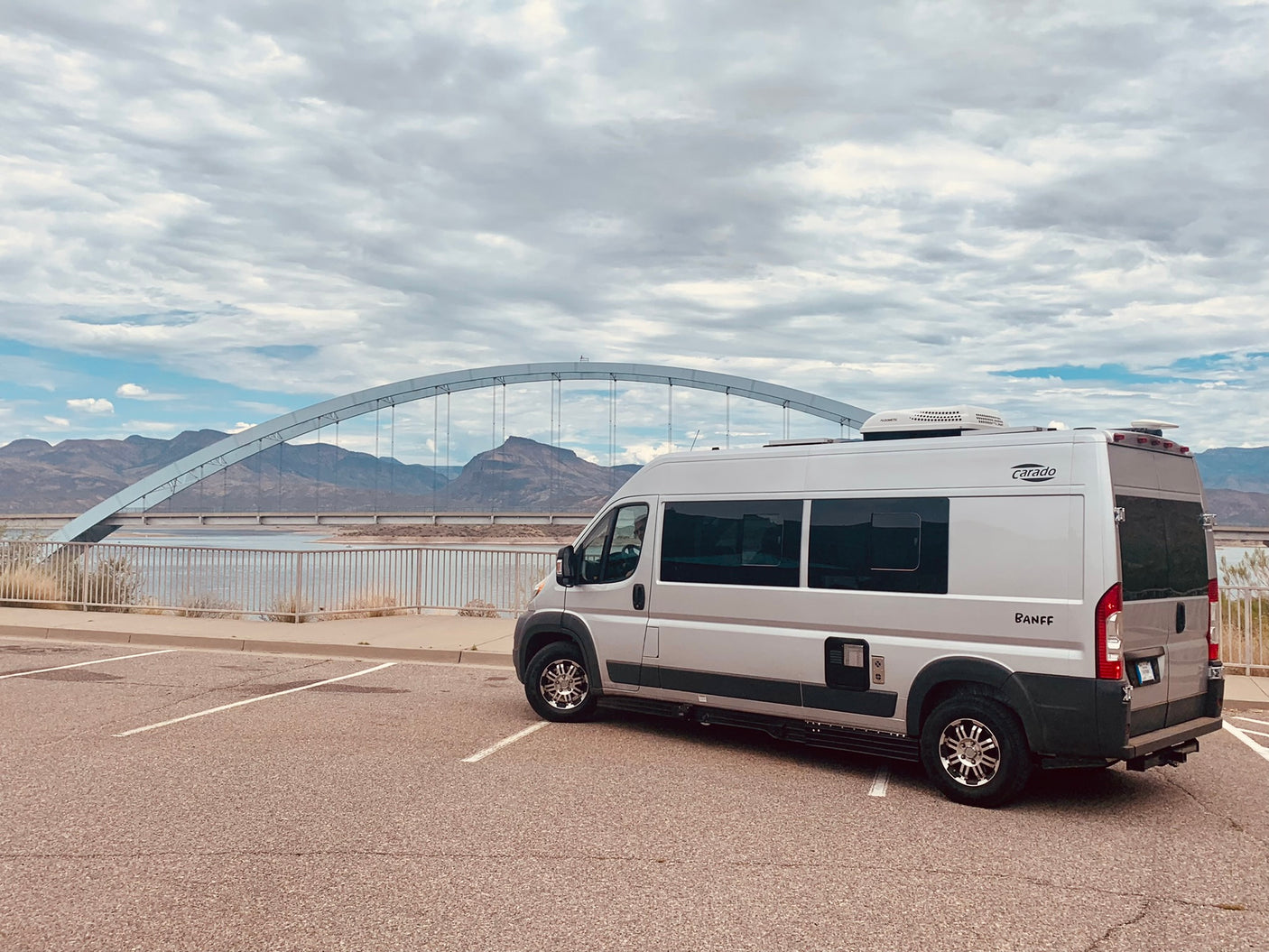 Silver Carado Banff RV rental parked by a scenic arch bridge over water, with mountains in the background under a cloudy sky.