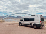 Silver Carado Banff RV rental parked by a scenic arch bridge over water, with mountains in the background under a cloudy sky.