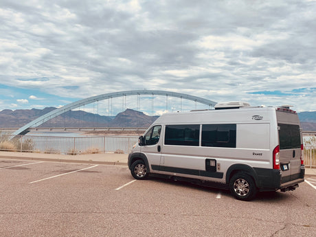 Silver Carado Banff RV rental parked by a scenic arch bridge over water, with mountains in the background under a cloudy sky.