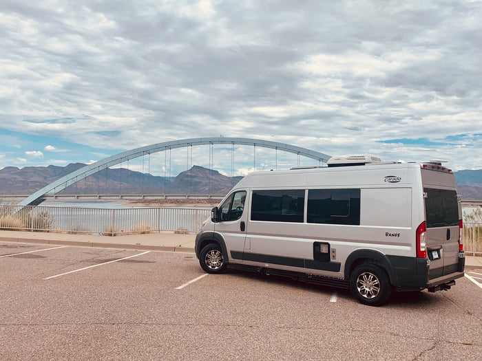 Silver Carado Banff RV rental parked by a scenic arch bridge over water, with mountains in the background under a cloudy sky.