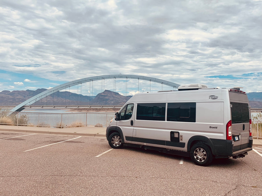 Silver Carado Banff RV rental parked by a scenic arch bridge over water, with mountains in the background under a cloudy sky.