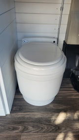 White portable toilet with closed lid on wood floor, installed in an rv rental vehicle with white slatted walls.