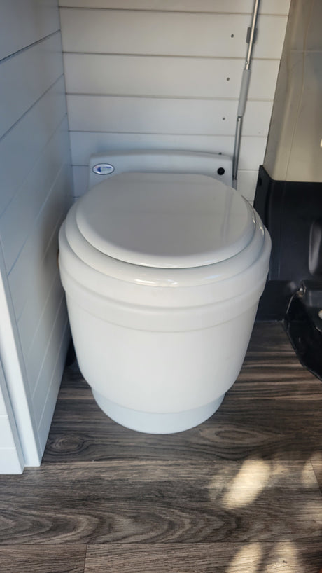 White portable toilet with closed lid on wood floor, installed in an rv rental vehicle with white slatted walls.