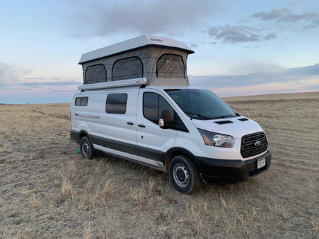 White camper van with pop-top tent in a dry field under cloudy skies, perfect for an RV rental getaway.