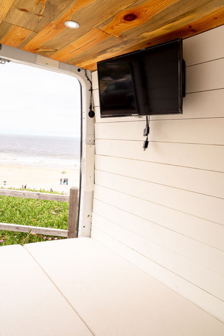 RV rental interior with a view of the beach, featuring a mounted TV, wooden ceiling, and comfy bed.