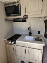 Compact RV rental kitchen featuring GE Profile microwave, 2-burner stovetop, sink, and light wood cabinets for efficient travel.