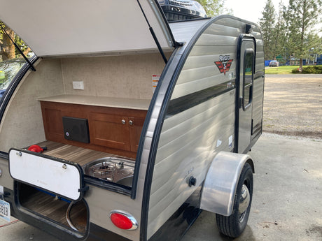 The open hatch reveals a compact outdoor kitchen with a sink, stovetop, and storage in this Retro Jr. travel trailer, perfect for an rv rental.