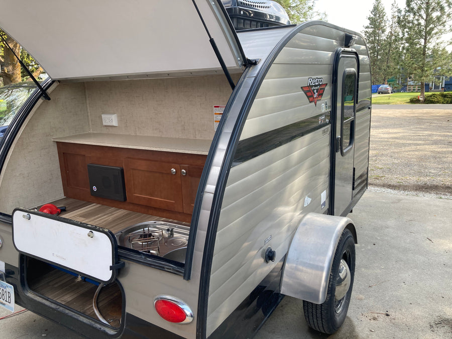 The open hatch reveals a compact outdoor kitchen with a sink, stovetop, and storage in this Retro Jr. travel trailer, perfect for an rv rental.