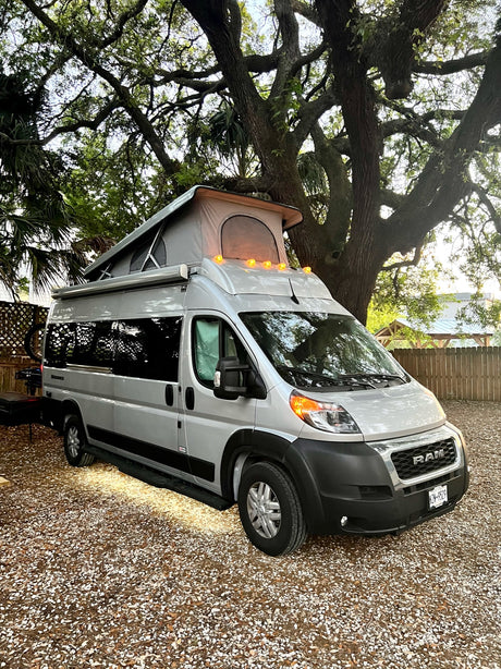This sleek silver pop-top camper van, an ideal RV rental, rests on gravel under a grand tree canopy.