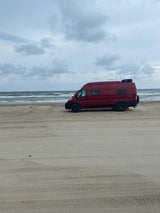 Red rv rental parked on a vast sandy beach with ocean waves under a cloudy sky, ready for adventure travel.