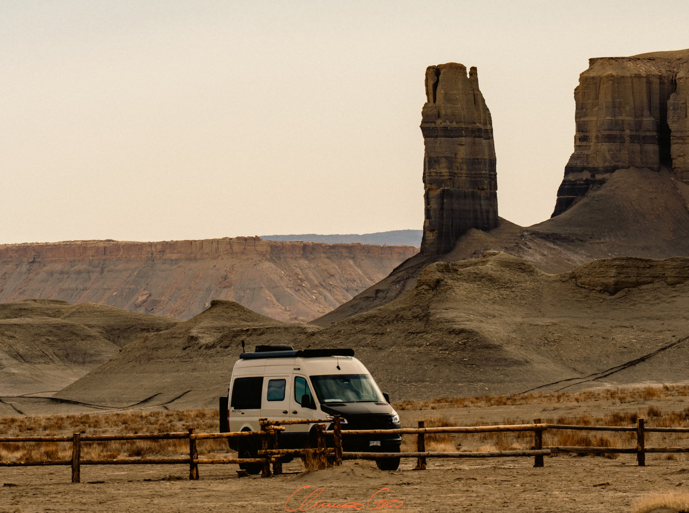 An RV rental camper van parked in a vast, arid landscape with impressive desert rock formations and a wooden fence.